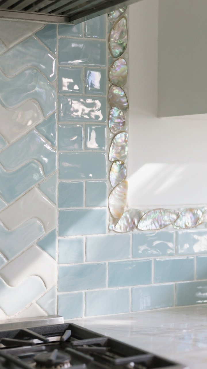Detail closeup of a coastal backsplash that sparkles like sea glass: glossy zellige-style tiles in pale blue-gray and off-white with slight color variation and watery waviness, light grout lines, a vertical stacked section near the range, and a subtle mother-of-pearl inlay border catching light; angled natural daylight bouncing off the glaze to create reflective highlights; tight crop showing tile surface, beveled edges, and shimmer.