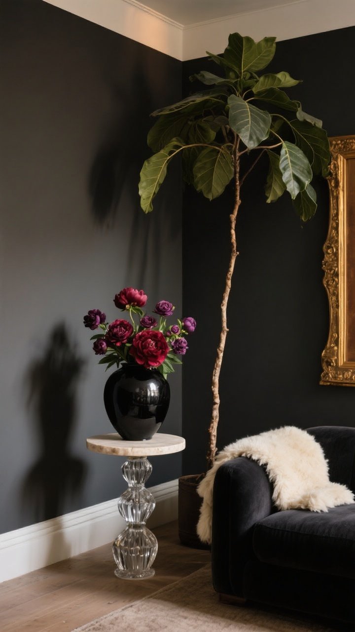Medium vignette from a corner angle — Botanical drama with contrast: a black ceramic vase holding burgundy peonies and plum ranunculus on a pale stone side table; a tall rubber tree adding architectural height beside matte charcoal walls; an antique gold frame catching warm light; a cream shearling throw draped over a dark sofa for yin-yang contrast; a single tall branch in a cut-crystal vase on the floor for sculptural impact; dim, warm lighting; no people.